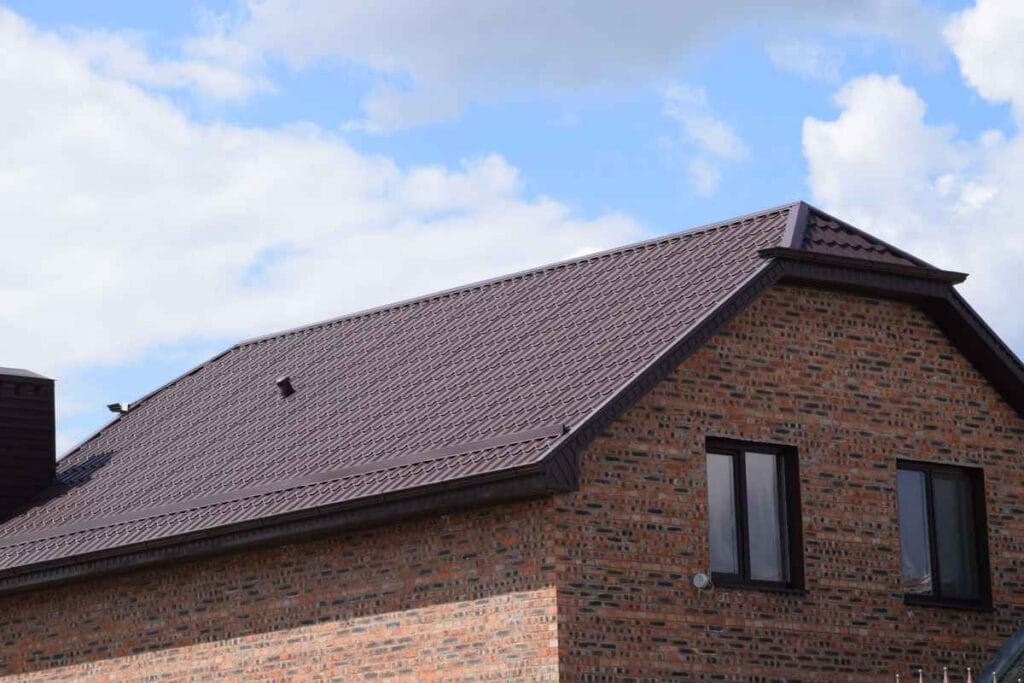 Brick house with a dark metal roof under a blue sky with clouds, crafted by a roofing contractor in Morris County, NJ. Two windows are visible on the side of the building, showcasing quality craftsmanship and attention to detail.