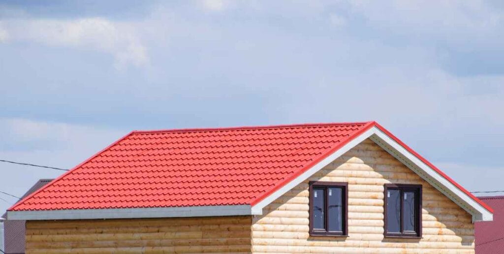 A wooden house with a bright red, peaked roof stands against the blue sky, reminiscent of roofing in Bergen County NJ. The log cabin design features two dark-framed windows on the upper level. Nearby, partial views of neighboring rooftops are visible, completing this picturesque scene.