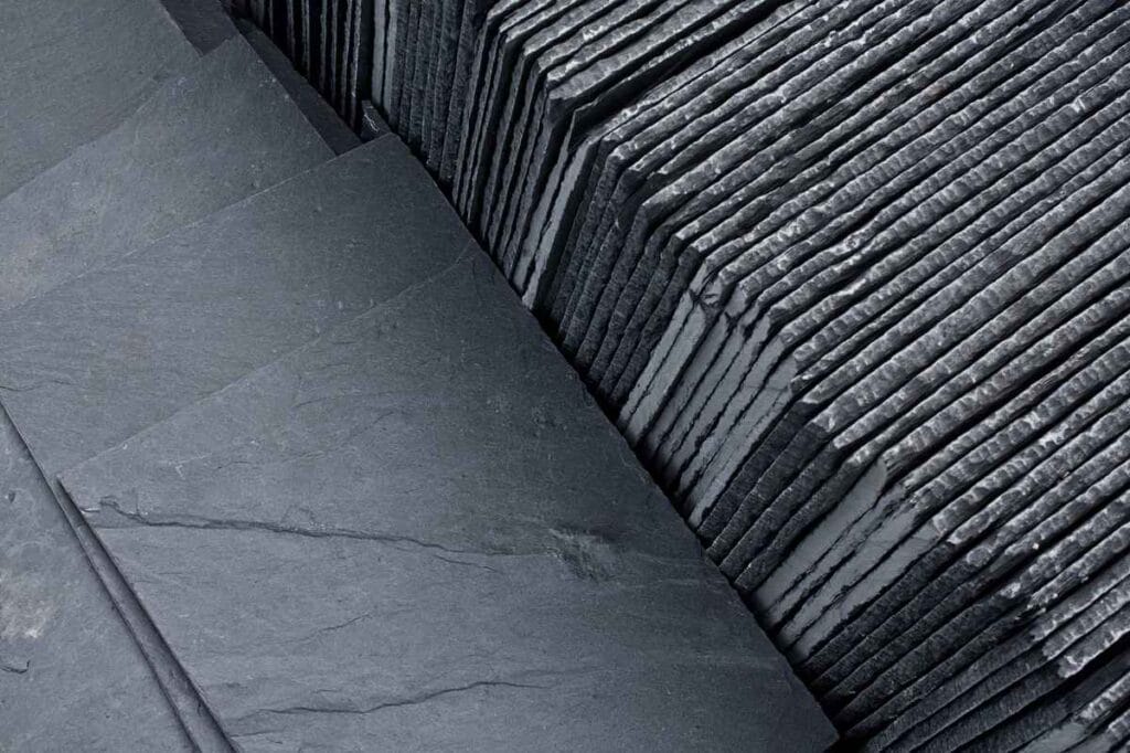 Stacked slate tiles arranged neatly, showcasing their textured surfaces and edges, are perfect for any roofing project in Passaic County, NJ. The tiles appear dark gray with natural striations, highlighting their rough and layered composition.