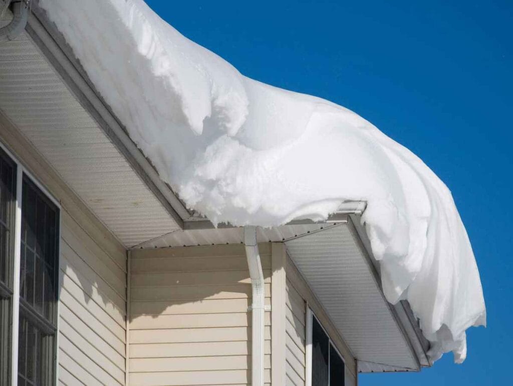 Snow is piled high on the roof of a light-colored house in Bergen County, NJ, creating an overhang. The clear blue sky contrasts with the bright white snow while a portion of the siding and windows is visible—a picturesque scene for any roofer in Bergen County to appreciate.
