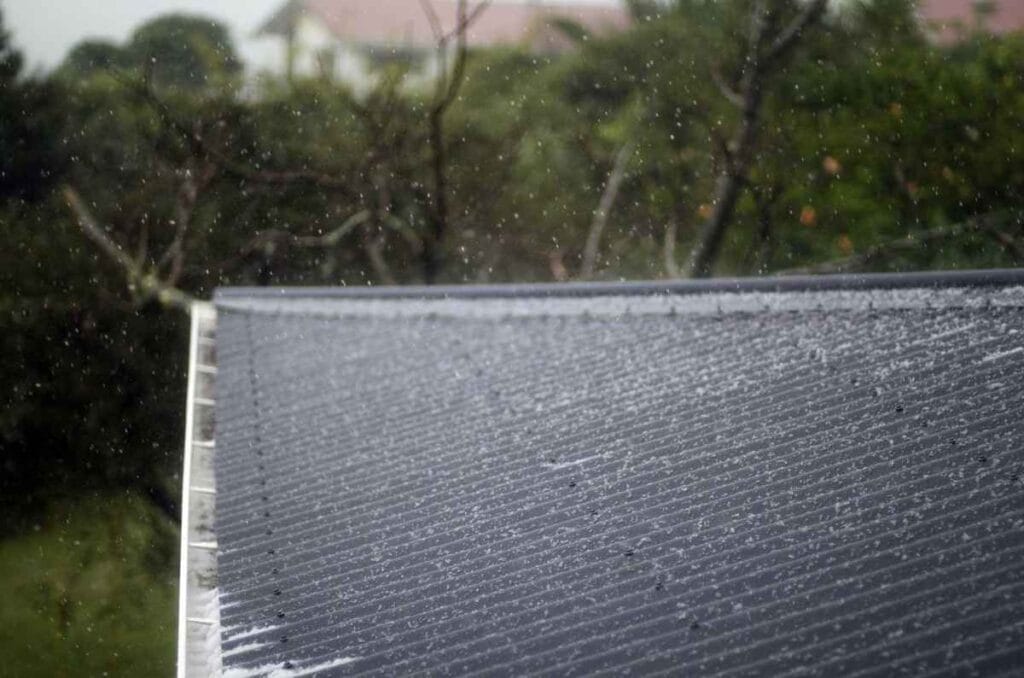Rain falls on a sloped corrugated metal roof, installed by a skilled roofer in Bergen County, NJ. Water droplets dance in the air amid trees and a blurred building in the background, creating a soft, rainy day atmosphere.