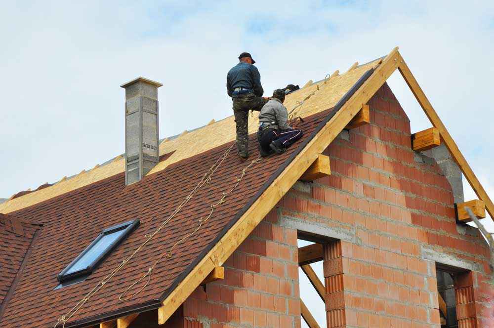 Two workers are on the roof of a partially constructed brick house, diligently placing wooden framework and shingles. The sky is clear over this project by a skilled roofing contractor in Morris County, NJ. The house boasts a chimney and a window nestled into the sloped roof.