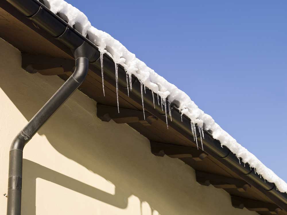 A house roof covered in snow and small icicles glistening under a clear blue sky, showcasing the expert work of a roofing contractor in Morris County NJ. A dark drainage pipe runs down the beige wall, completing this picturesque winter scene.