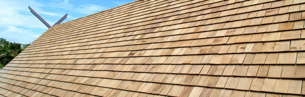 A wooden shingle roof with a steep slope, expertly crafted by a roof company in Passaic County NJ, is shown under a blue sky. The shingles are uniformly laid, creating a neat pattern. Palm trees are visible in the background on the left side.