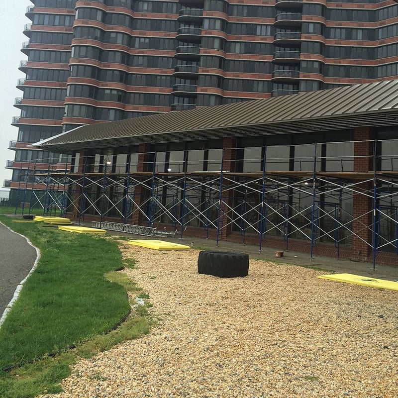 A large brick building with scaffolding on the lower floor suggests a roofing contractor in Morris County, NJ, might be at work. A row of windows shows off its wavy design. In the foreground, a gravel path is scattered with a large black tire and some yellow boards.