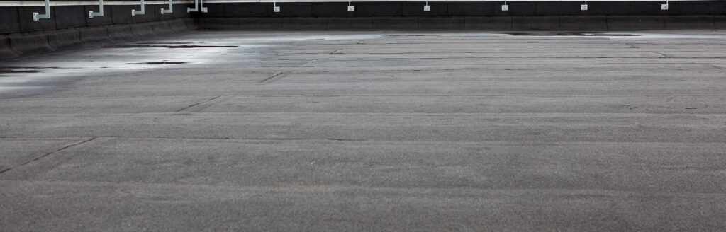 The flat rooftop, likely maintained by a reputable roof company in Passaic County NJ, features a gray surface with faint markings and slightly wet areas. Bordered by a low railing, the overcast sky casts a uniform hue across the scene.