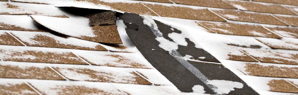 Close-up of a damaged roof in Bergen County, NJ, with brown shingles partially covered in snow. Several shingles are lifted, exposing the underlayment beneath. Snow has accumulated in small patches across the surface.