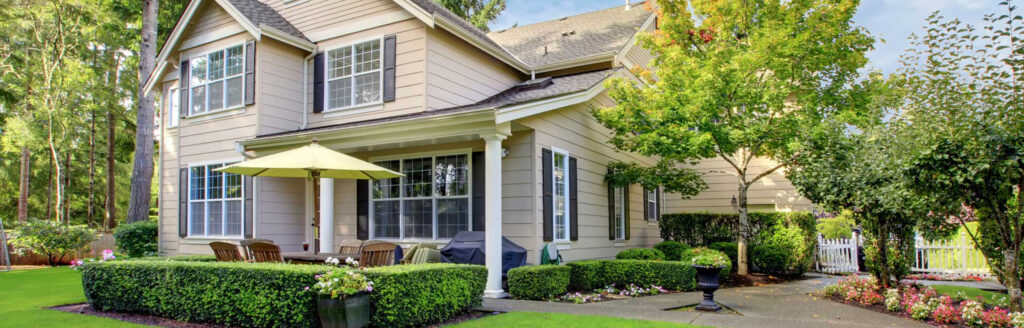 A two-story beige house with white trim, large windows, and a gabled roof showcases expert craftsmanship by a roofing contractor in Morris County NJ. The front yard features a patio with a dining set under a green umbrella, surrounded by manicured bushes and lush trees. A white fence is in the background.