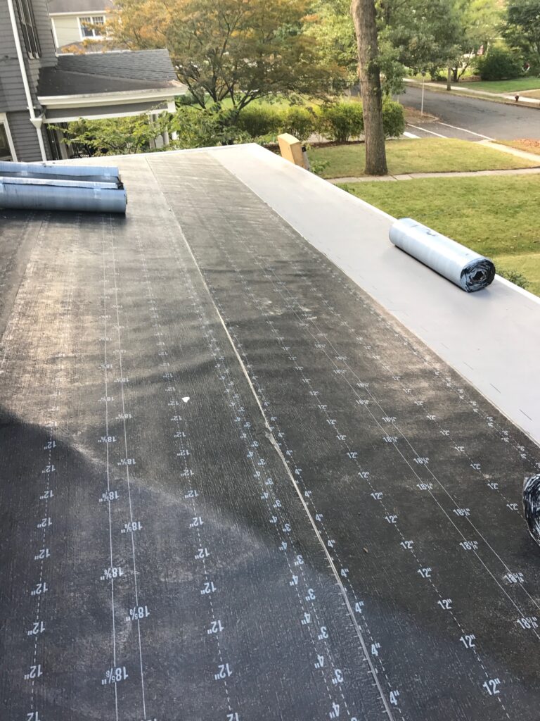 A flat roof under construction is covered with a black waterproofing membrane, with rolled-up materials laid out, courtesy of a roofing contractor in Morris County, NJ. The scene features trees, a street, and grass in the surroundings.