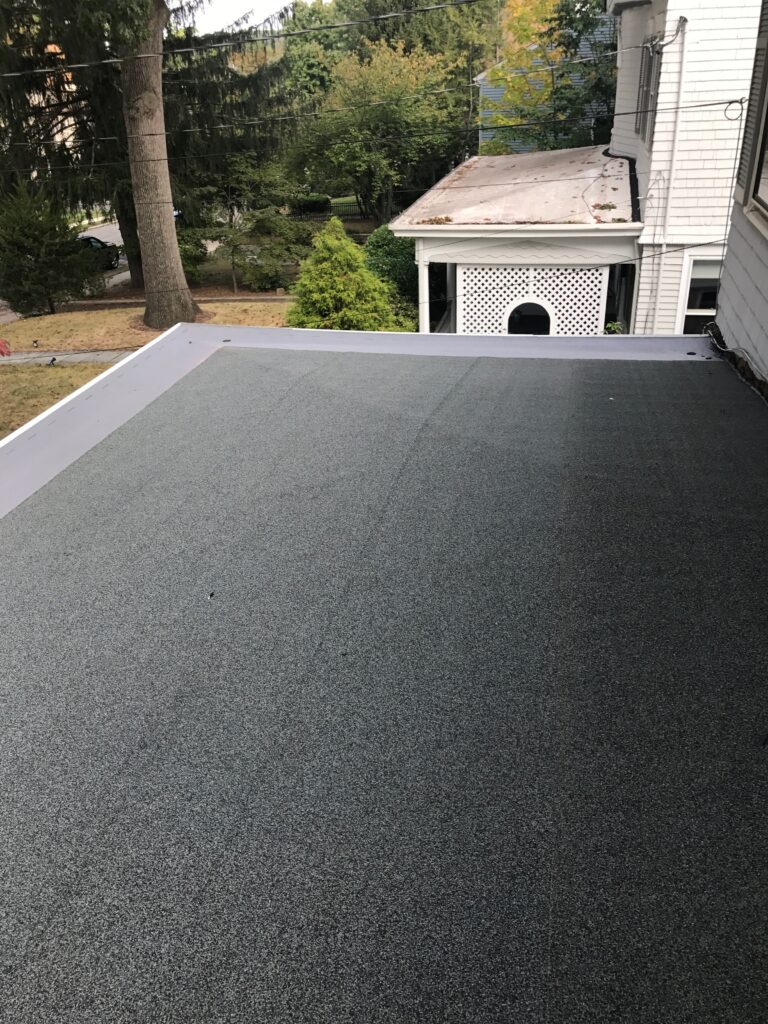 Flat rooftop with a tarred surface in a residential area. A tree and a neighboring building with white siding are visible in the background. The scene suggests an overcast day, typical for roofing in Bergen County, NJ, within a suburban setting.