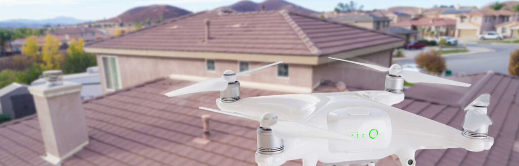 A white drone hovers in the foreground above a residential neighborhood, showcasing houses with brown roofs. The scene, perfect for a roofing contractor in Morris County NJ, also captures distant hills and a partly cloudy sky.
