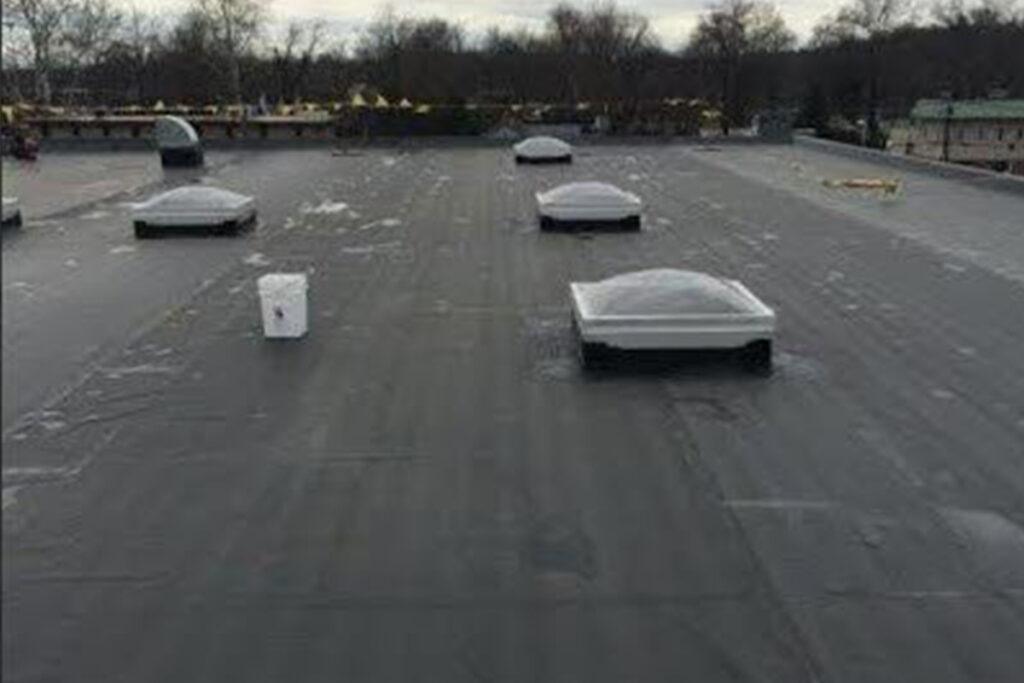 A flat rooftop with several dome-shaped skylights and a white vent, surrounded by bare trees and some buildings in the background on a cloudy day.