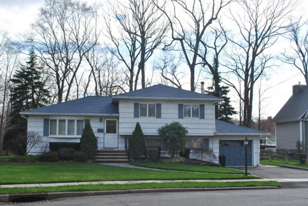 A split-level suburban house with light siding, blue shutters, and a blue garage door, surrounded by a manicured lawn and trimmed bushes. Leafless trees stand tall in the background against a partly cloudy sky.