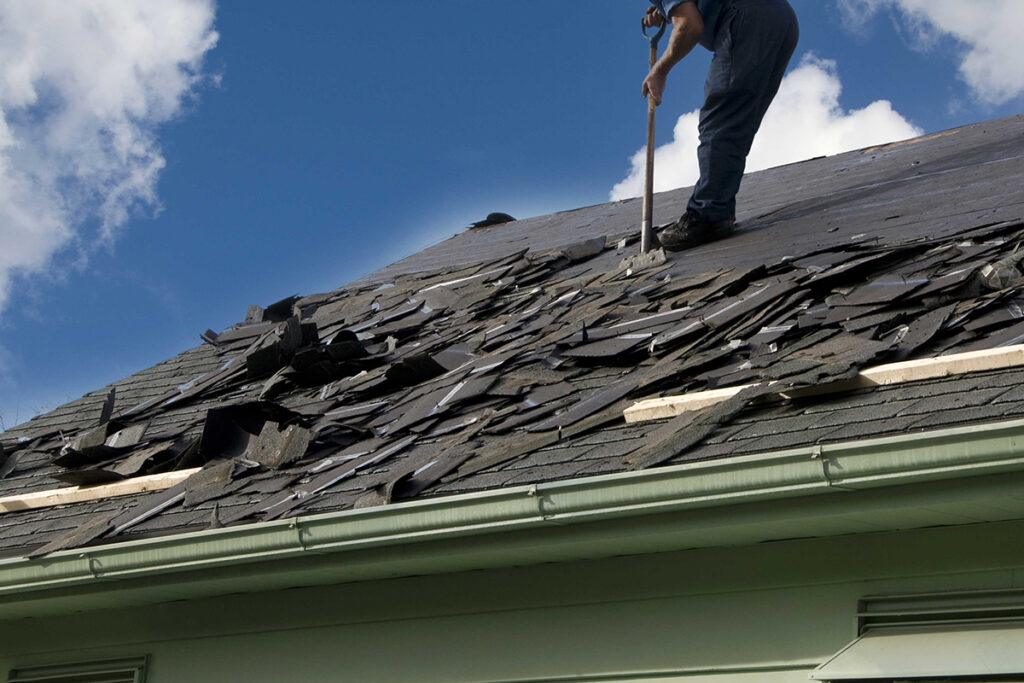 A worker uses a tool to remove old, damaged shingles from a house roof under a blue sky with clouds. Piles of shingles are scattered across the rooftop.