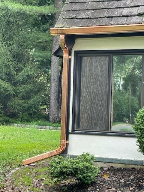 A copper downspout runs from the roof to the ground along the corner of a house with large windows, next to green grass, shrubs, and tall evergreen trees in the background.