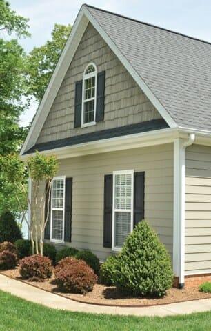 A beige house with white trim and black shutters, featuring a peaked roof, upper arched window, and neatly landscaped shrubs and small trees in front.