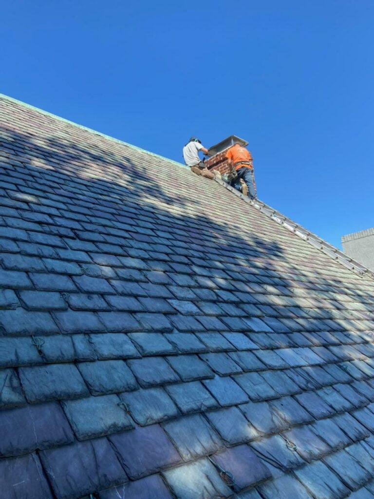 Two workers stand on a ladder near the top of a steep, slate-tiled roof, working on a chimney under a clear blue sky. Shadows from nearby trees fall across the roof.