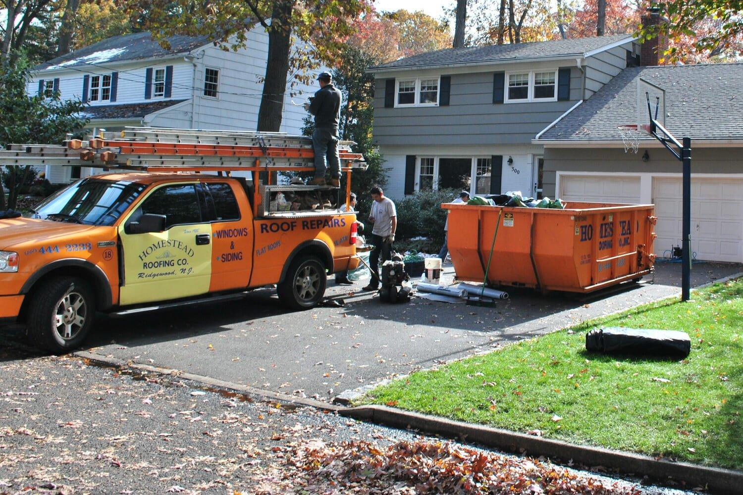 An orange roofing company truck is parked in front of a house, with workers and equipment nearby. A large dumpster sits in the driveway, and leaves are scattered on the ground. A basketball hoop is visible by the garage.