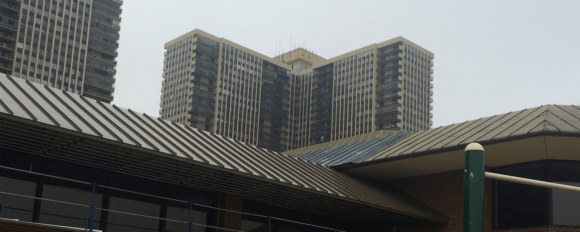 Tall high-rise buildings with many windows and balconies rise behind lower, angular-roofed structures in the foreground under a cloudy sky.