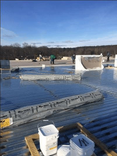 Workers installing roofing material on a large, flat commercial roof under a clear blue sky, with construction supplies and tools scattered around. Trees and a forested area are visible in the background.
