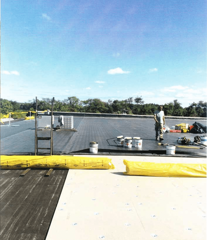 A worker stands on a flat rooftop under a blue sky, surrounded by construction materials, buckets, and tools. A ladder and yellow insulation material are also visible on the roof. Trees are in the background.