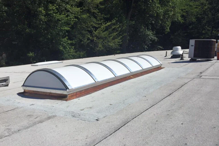 A long, curved skylight is installed on a flat, gray roof with trees in the background and HVAC units nearby.