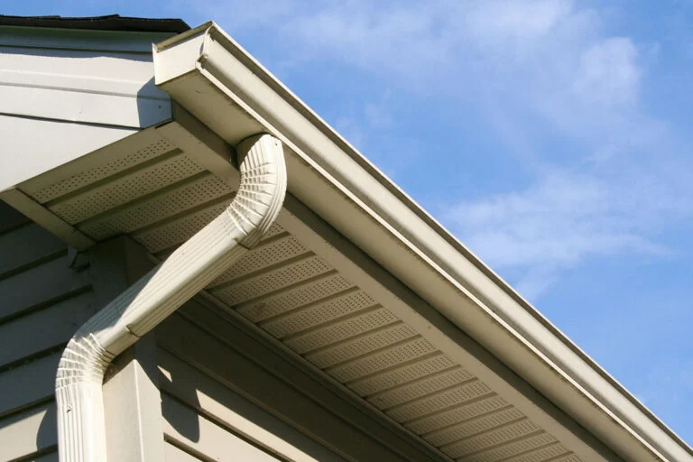 Close-up view of a house’s roof edge with a white rain gutter and downspout against a blue sky with some clouds. The exterior siding and soffit vents are also visible.