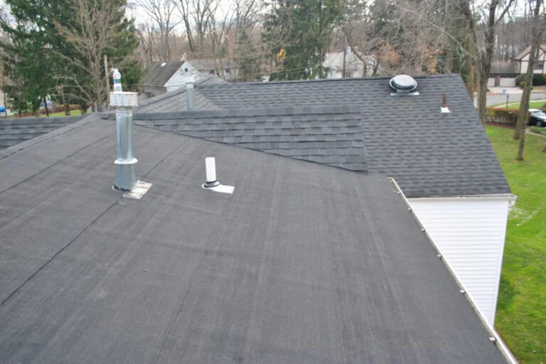 A house roof with black asphalt shingles and a flat black section featuring several metal vents and pipes, surrounded by trees and neighboring houses in the background.
