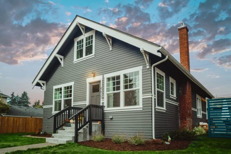 A gray house with white trim, a brick chimney, and a small front porch with steps, set against a blue sky with scattered clouds at sunset. The lawn and landscaping are neatly maintained.