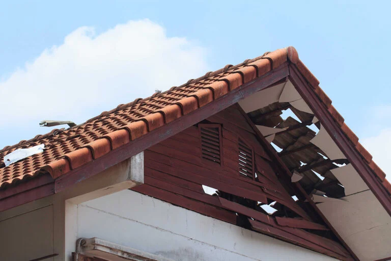 The image shows a house roof with red tiles, some of which are broken or missing, revealing damage to the underlying structure against a blue sky with clouds.