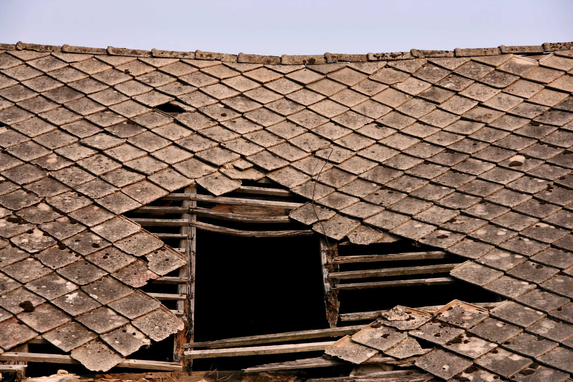 A close-up of an old, damaged roof with broken and missing tiles, revealing wooden beams and a dark, rectangular hole beneath. The remaining tiles are weathered and uneven.