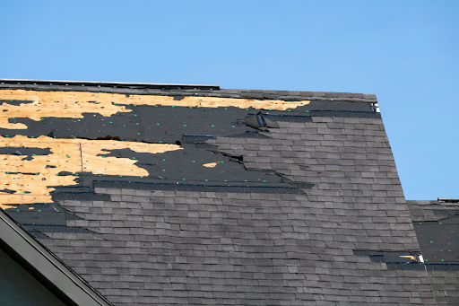 A house roof with missing and damaged shingles, exposing the underlying wooden structure, likely caused by storm or wind damage, under a clear blue sky.